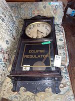 Full view of the Eclipse Regulator clock showing the entire wooden case, glass panel with text, pendulum, and winding key on the front glass.