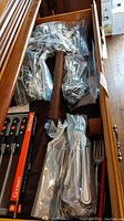 Drawer filled with various mixed pattern cutlery pieces in plastic bags, including forks and spoons, and a boxed set of steak knives.