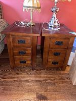 Photo of two identical wooden end tables, each with three drawers and black metal handles, positioned side by side on a wooden floor against a red wall, with lamps on top.