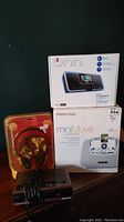Boxed Onn portable 2.0 speaker, Memorex miniMove iPod dock, Panasonic clock radio with wired headphones in plastic clamshell case, all placed on wooden surface against dark background.