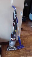 Full shot of Shark Professional Powered Lift-Away vacuum cleaner and Swiffer WetJet mop standing next to wall on wooden floor.