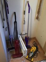 Photo showing multiple vacuum cleaners including purple stick vacuum, red upright vacuum, white floor grate, black upright vacuum, and yellow handheld vacuum