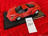 Photo of a red Ferrari Enzo car model placed on a black display base with Ferrari branding, photographed on a red fabric background.