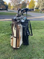 Two golf bags standing on grass outdoors, each filled with golf clubs including drivers, irons, and putters. The bags are tan/white and black in color respectively.