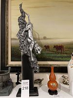 Photo showing the silver-tone jaguar figure sculpture climbing down a textured vertical base, mounted on a smooth black rectangular stand with a pastoral painting and other decorative items in the background.