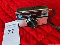 Front view of a Kodak Instamatic 155x camera on a red quilted fabric surface with a wrist strap and MaxSold auction lot tag numbered 77