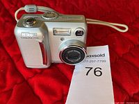 Silver Nikon Coolpix 775 digital camera on red background with wrist strap visible, front side showing lens and flash.