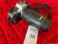 Front and side view of silver and black Pentax film camera with attached black telephoto zoom lens and camera strap on red background.