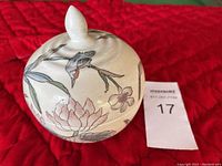 Closed view of round porcelain trinket box with lid showing floral and bird painted design on cream background, with finial handle on lid.