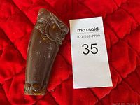 Photo showing the full carved horn sculpture from the side with carved buffalo head detail, placed on red quilted fabric next to paper tag showing lot number.