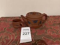 Photo showing the purple clay teapot and matching cup with hand-painted blue floral and butterfly designs placed on a patterned fabric surface.