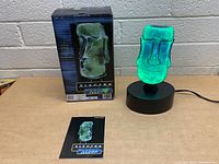 Electra Mini Moai Plasma Light illuminated in green on black round base with original retail box and instruction manual placed in front.