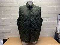 Front view of green diamond-quilted vest with full zipper and stand collar on mannequin against beige brick wall.