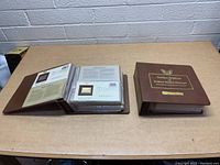 Two hardcover books on table, showing closed and open book with gold replica stamp pages inside.