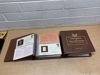 Two brown hardcover books titled 'Golden Replicas of United States Stamps' with pages of first day cover envelopes containing gold replica stamps inside plastic sleeves visible on table surface in front of white brick wall.