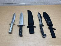 Three fixed-blade knives laid out alongside their black nylon sheaths on a wood surface, showing full length and handle details.