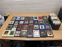 Lot of 38 assorted 8 track tapes laid out on a table, showing multiple different album covers and colors of tape cases. Mainly black cases with some in blue, white, or red.