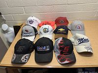 Photo showing twelve assorted hats in various colors and logo designs on a table against a wall.