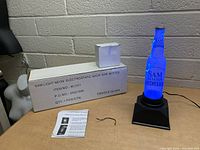 Photo showing the blue neon-lit Sam Adams bottle on black base, with original box and instruction manual on table.