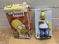 The Simpsons Homer tin action toy next to its original colorful box showing front artwork with Homer Simpson and logos.