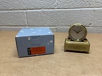 Photo of vintage Reuge Swiss alarm clock with decorative gold-tone metal rectangular base and round clock face, placed next to original blue box.