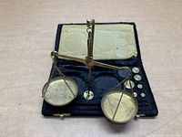 Top down view of vintage brass balance scale with pans and attached chains inside an open black velvet-lined molded case with round brass weights arranged.