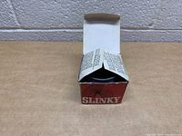 Top view of the vintage slinky inside the partially opened box, showing the black coil and inner flaps of the box.