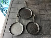 Photo showing all three cast iron frying pans arranged on a black grid surface, visible different textures and shape of handles with holes for hanging.