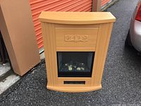 Front view of modern electric fireplace with light wood finish and decorative carved floral motif above glass window.