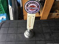 Full view of the handheld thermometer standing on a metallic base with a vertical scale visible and the round Harley Davidson Genuine Motor Oil sign on top.