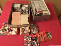 Wide view of two large boxes filled with approximately 3000 sports cards and several smaller stacks of cards laid out showing various players and card faces.