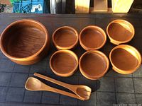 Photo shows the complete set with one large master bowl, six smaller serving bowls, and two wooden salad utensils placed on a black surface.