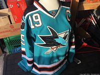 Full front and side view of teal hockey jersey on mannequin showing San Jose Sharks logo, number 19, and captain 'C' patch.