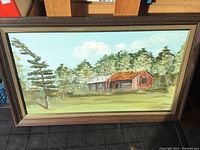 Framed oil on board painting showing a red and gray barn surrounded by trees and grass.