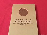 Tan presentation pouch with circular window showcasing the Silver Jubilee commemorative coin, labeled with text commemorating Queen Elizabeth II's Silver Jubilee from 1952-1977.