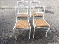 Four grey painted wooden dining chairs with upholstered seats and carved top backrests, arranged outdoors on pavement.