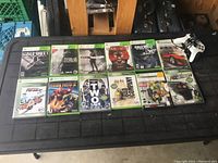 Photo showing all 11 Xbox 360 game cases and one white wired Xbox 360 controller laid out on a black mat.