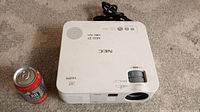 Top view of white NEC projector showing label VE281X and HDMI logo, power cable coiled behind, size comparison with standard soda can.