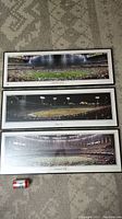 Three framed panoramic stadium photos by Rob Arra laid on carpet with soda can for scale, showing wide shots of large stadiums filled with crowds.