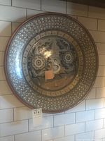 Front view of round decorative platter hung against a tiled wall, showing floral patterns and geometric border.