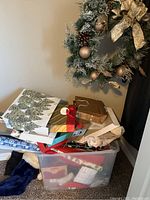 Plastic bin filled with assorted Christmas gift bags of different sizes and designs, next to a decorative Christmas wreath with gold ornaments and bow hung on wall.