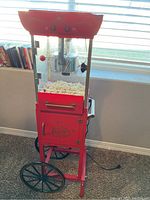 Full front view of red popcorn maker with glass enclosure filled with popcorn on a wheeled cart near window.