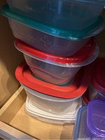 Stack of plastic food storage containers with assorted colored lids inside a kitchen cabinet.