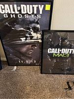 Two framed Call of Duty posters side by side on carpeted floor, one labeled Ghosts and dated 11.5.13, the other MW3 with two soldiers.