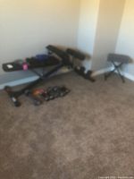 Photo of the exercise bench alongside assorted free weights on the carpeted floor of a room corner.