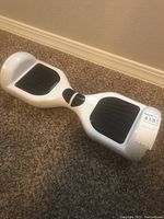 White hoverboard resting on carpet, displaying the top and side with black footpads and a label on the side.