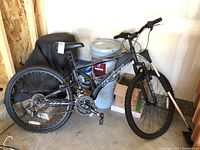 Full view of the black Hyper Havoc FS mountain bike with visible 26-inch wheels, full suspension frame, and components. Bike is positioned indoors near paint buckets and boxes.