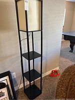 Tall black metal floor lamp shelf with three square shelves and power cord visible, photographed standing upright near wall and furniture.