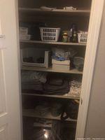 View of closet shelves showing white plastic baskets holding cleaning supplies, bottles, and wipes, along with folded linens and boots in a plastic shoe organizer.