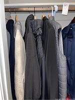 Photo shows seven men's outerwear pieces hanging on wooden hangers in a closet, including jackets and vests of various colors and materials.